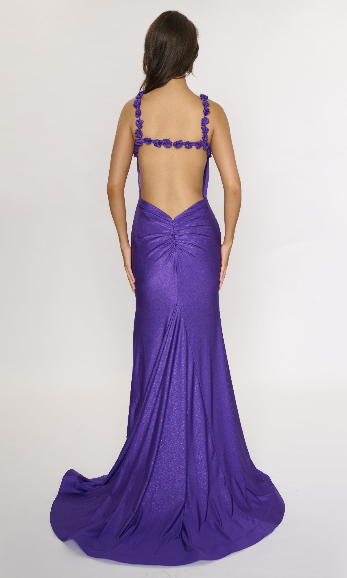 A woman stands with her back to the camera against a plain light background, wearing the floor-length Nina Canacci 5304 formal dress featuring floral straps and a backless design.